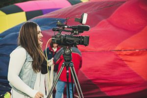 A female videographer operates a camera before a colorful hot air balloon backdrop.