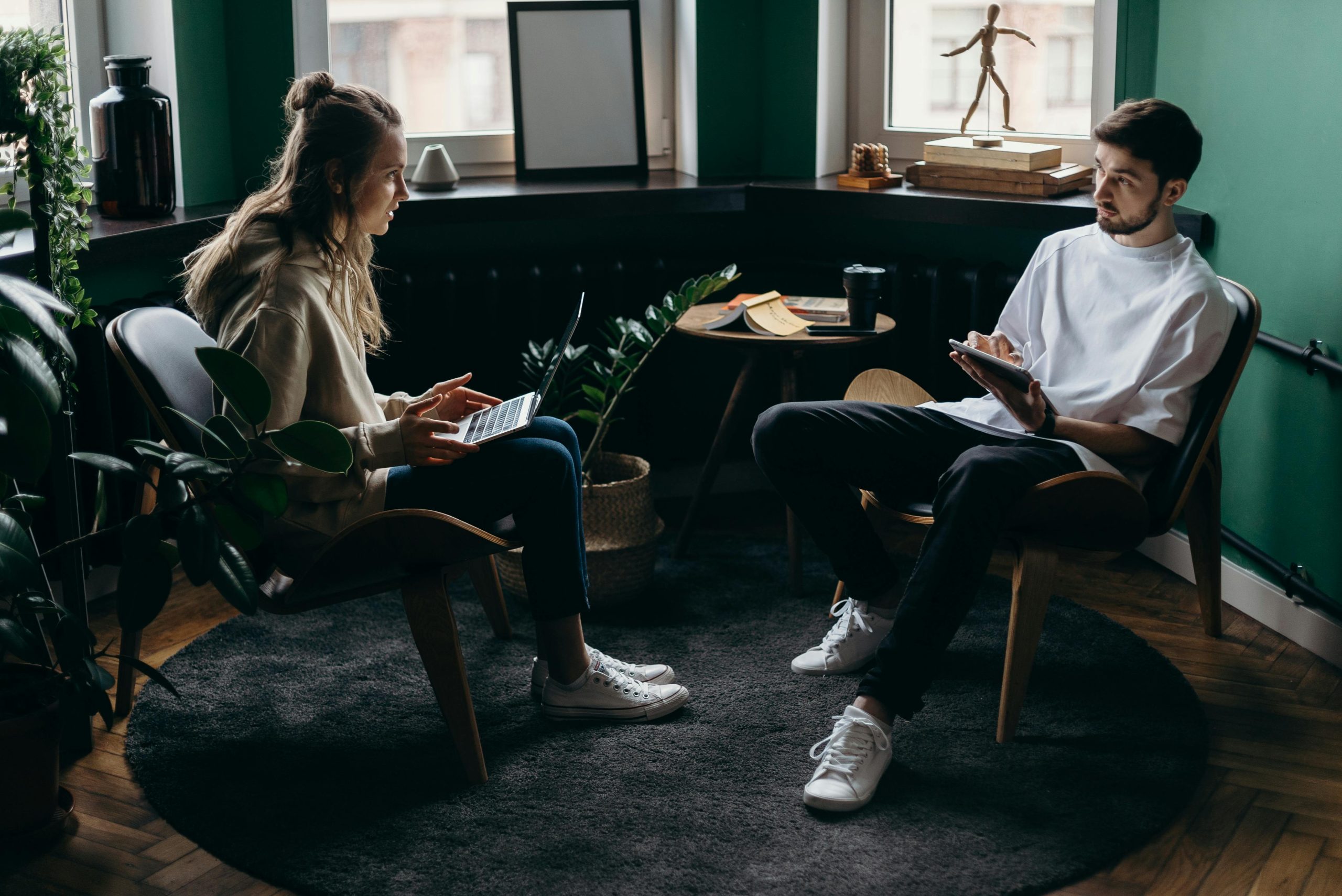 A man and woman engaged in conversation at a stylish, plant-filled home office with modern furniture and digital devices.