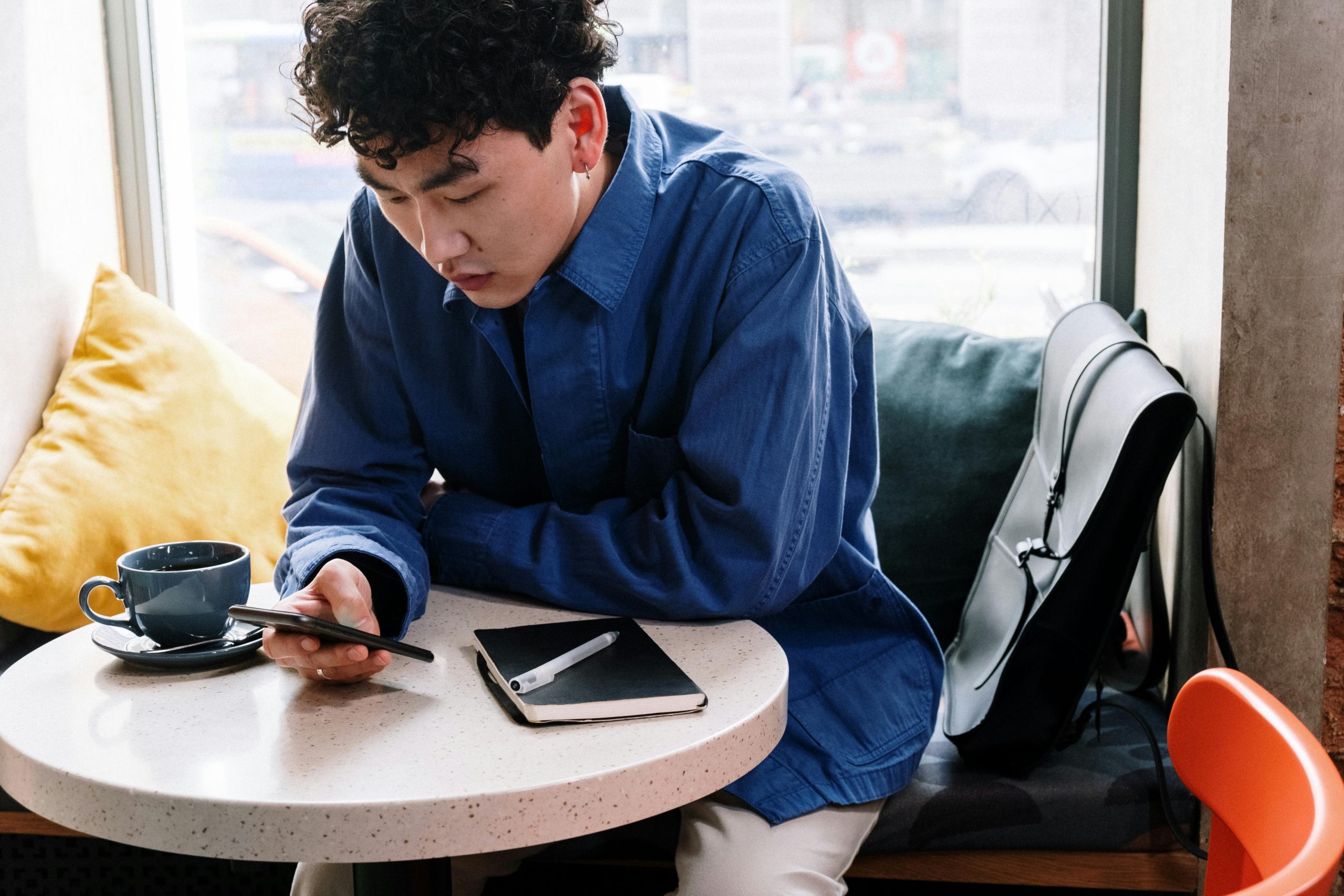 Young man in a cafe using a smartphone, with coffee and notebook on the table.